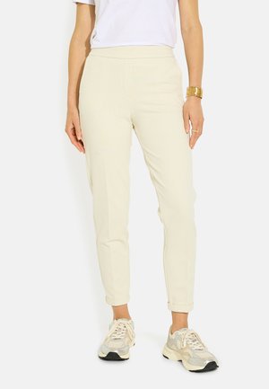 Mujer con pantalones beige entallados, zapatillas blancas, camiseta blanca y pulsera dorada de pie frente a un fondo blanco.