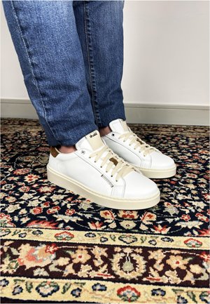 Persona che indossa sneakers bianche in pelle e jeans blu in piedi su un tappeto con motivi floreali e geometrici.