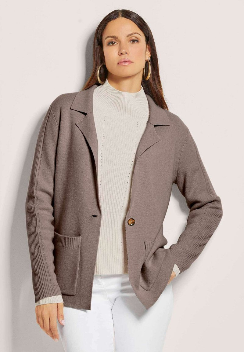 Braune Strickjacke mit übergroßen Taschen, Knopfverschluss und gerippten Ärmeln, kombiniert mit einem cremefarbenen Rollkragenpullover.