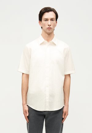 Noor mees, kellel on lühikesed tumejuuksed, kannab lihtsat valget lühikese varrukaga nööpidega särki ja tumedaid pükse, seistes heleda tausta ees.