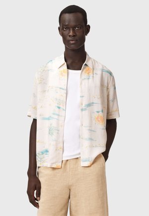 Mand iført beige shorts, hvid t-shirt og kortærmet skjorte med pastelfarvet sol- og landskabsprint, stående mod en ensfarvet baggrund.