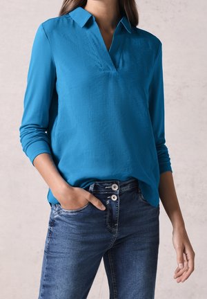 Femme portant un chemisier bleu à manches longues avec un col en V et un jean bleu foncé, une main dans la poche, debout devant un fond neutre.