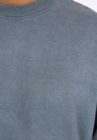 Baumwoll-T-Shirt in gedämpftem Blau mit Rundhalsausschnitt, das ein dezentes, geprägtes Logo auf der Brust und eine glatte, leicht strukturierte Oberfläche aufweist.