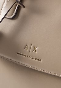 Λεπτομέρεια μπεζ δερμάτινης τσάντας που δείχνει το ανάγλυφο χρυσό λογότυπο Armani Exchange με τα αρχικά A/X και διακοσμητικό γαζί κοντά στην πάνω γωνία με θηλιά για λουράκι.