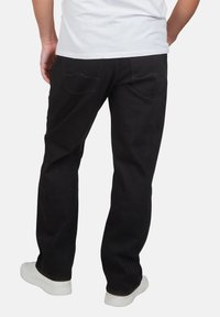 Hombre vestido con jeans rectos negros, zapatillas blancas y una camiseta blanca, de pie con las manos detrás de la espalda sobre un fondo blanco.
