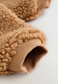 Karamelli fleece kangas, millel on lokkis tekstuur, kontrasteerib sujuva ribaga pruuni mansetiga, tuues esile erinevad materjalid ja tunnetuslikud omadused.