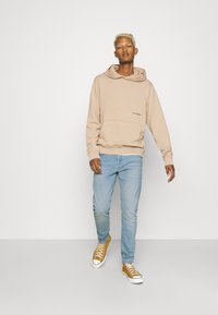Bež hoodie s kengurubskom džepom, nosi se s svijetloplavim trapericama. Model nosi smeđe tenisice s bijelim đonom, prikazujući ležeran izgled.