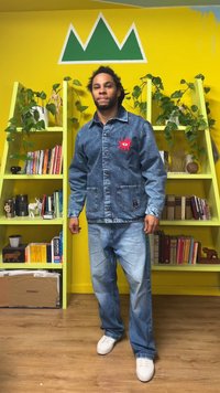 Džinsinė striukė su priekinėmis kišenėmis, puošta raudonu siuvinėtu raštu, derinama su šviesiai mėlynais džinsais ir baltais sportbačiais. Ryški geltona fono spalva.