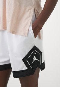 Calções atléticos Jordan em poliéster branco com detalhes em preto. Possui um emblema em forma de diamante e logo. Tecido texturizado com bolsos laterais.