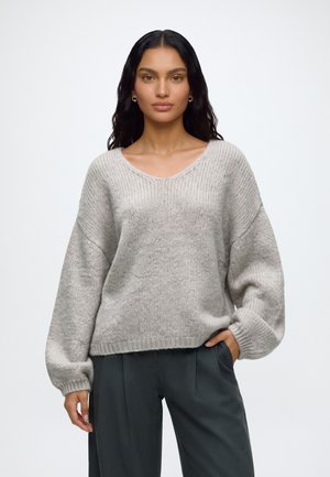 Mujer con cabello largo y oscuro que lleva pendientes de aro dorados, un suéter de punto gris claro y pantalones gris oscuro con una mano en el bolsillo.