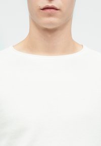 Młody dorosły ubrany w gładką, białą koszulkę z okrągłym dekoltem, widoczny od podbródka do górnej części klatki piersiowej na neutralnym tle.