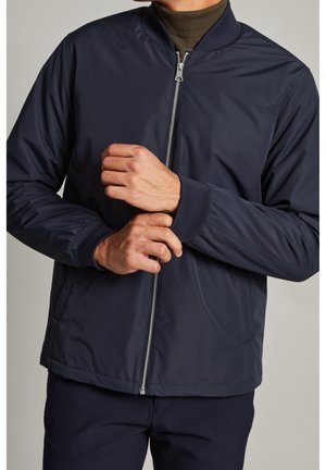 Homme portant une veste bomber bleu marine avec fermeture éclair devant sur un col roulé olive, ajustant le poignet de la manche gauche avec la main droite.