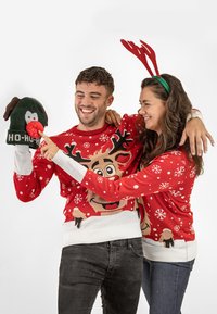 Red holiday sweaters with reindeer designs, white cuffs, and snowflake patterns. Man wears a green hat with a brown mitt and red pom-pom.