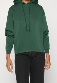 Grön huvtröja i mjukt material, med dragsnodd, avslappnad passform och ribbade ärmslut; parat med svarta jeans.