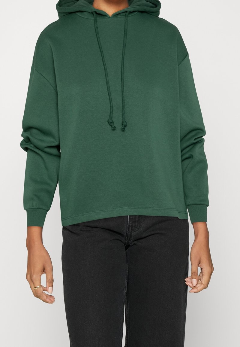 Grön huvtröja i mjukt material, med dragsnodd, avslappnad passform och ribbade ärmslut; parat med svarta jeans.