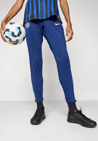 Blå treningsbukser med en avsmalnende design, ribbede mansjetter og et Nike-logo; kombinert med sorte fotballsko og holder en mønstret fotball.