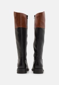 Bottes noires en cuir montantes jusqu'au genou avec un revers en cuir marron, dotées d'une fermeture éclair sur le côté et d'un petit talon empilé. Texture lisse et design épuré.