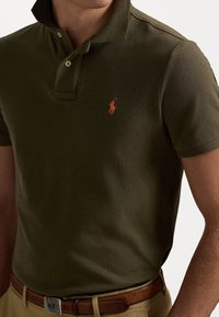 Mann trägt ein olivgrünes Poloshirt mit orangefarbenem gesticktem Logo, kombiniert mit beigen Hosen und einem braunen Ledergürtel mit silberner Schnalle.