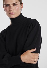 Pull noir en tricot avec un col montant, des poignets côtelés et des épaules légèrement bouffantes. Texture lisse et silhouette ajustée.