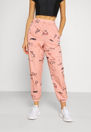 Osoba nosząca różowe joggery z wysokim stanem z czarnym i czerwonym ręcznie pisanym nadrukiem oraz białe trampki, w połączeniu z czarnym, krótkim topem.