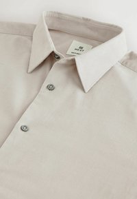 Helle beige Hemdbluse aus strukturiertem Stoff, mit spitzem Kragen und grauen Knöpfen vorne. Das Etikett trägt die Aufschrift "NEXT RELAXED."