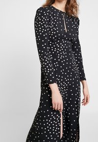 Svart klänning med prickar och långa ärmar, med en splitt i kjolen och nyckelhålsdetalj vid halsringningen. Slät tygtextur.