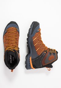 Bottes de randonnée montantes avec un schéma de couleurs noir et orange, en tissu en maille, semelle en caoutchouc et embout renforcé pour la durabilité.