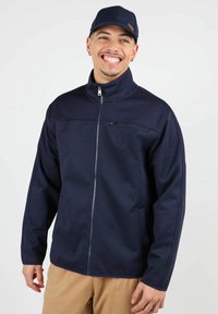 Veste zippée bleu marine en tissu texturé avec un col haut et des poches avant. Présente un petit logo sur la poitrine.