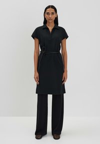 Robe noire à col avec des manches courtes, ceinturée à la taille. Portée par-dessus un pantalon noir ample. Texture de tissu lisse, design minimaliste.