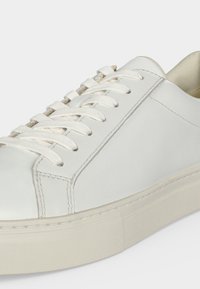 Witte lage sneaker met witte veters, glad leren bovenwerk en dikke gebroken witte rubberen zool, in close-up getoond.