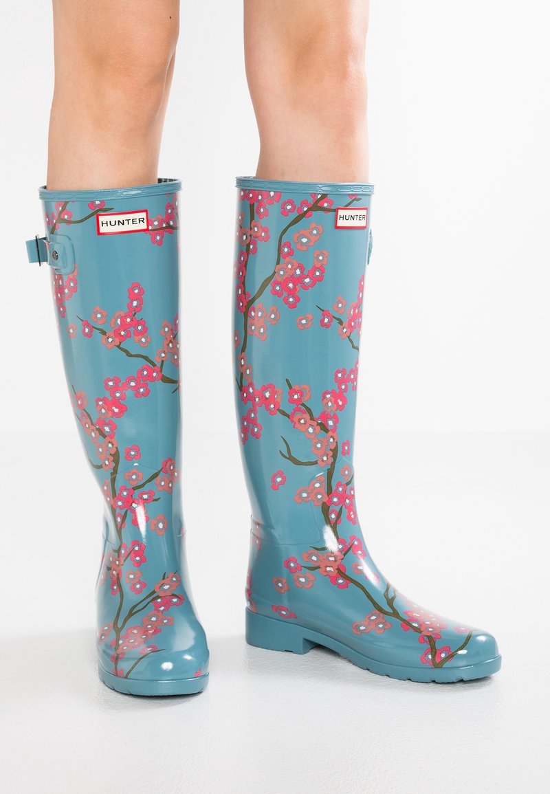 Botas de lluvia de goma azules con un diseño floral que presenta flores rosas y ramas marrones. Las botas tienen un tacón bajo y un hebilla ajustable.