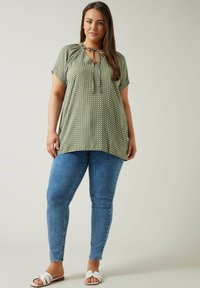 Grüne Bluse mit Polka-Dots, kurzen Ärmeln, Krawattenhals und lockerer Passform, kombiniert mit blauen Skinny-Jeans und weißen Sandalen.