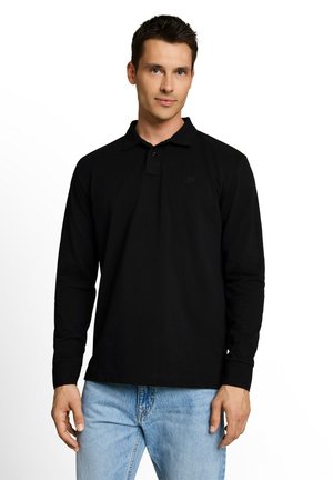 Homme debout portant un polo à manches longues noir et un jean bleu clair sur fond blanc.