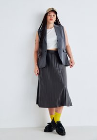 Blazer grigio senza maniche sopra a una maglietta bianca corta, abbinato a una gonna plissettata grigio scuro. Le scarpe nere e i calzini gialli brillanti completano l'outfit.