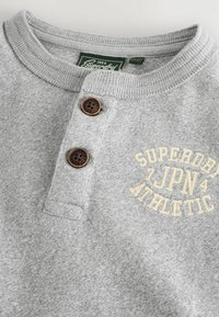 Grauer Baumwollpullover im Henley-Design mit zwei braunen Knöpfen und aufgesticktem "SUPERDRY JPN ATHLETIC" in Creme auf der Brust.
