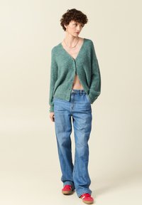 Grön stickad cardigan med V-ringning och knäppa, tillsammans med högmidjade blå jeans och röda sneakers. Mjuk textur, avslappnad passform.