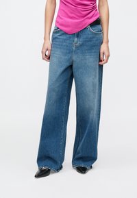 Femeie purtând blugi de denim largi albaștri, top roz, pantofi negri cu vârf ascuțit, stând pe un fundal alb.