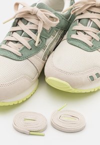 Ljust beige och gröna sneakers med beige snören uppradade, placerade bredvid två hoprullade beige ersättningssnören på en vit yta.