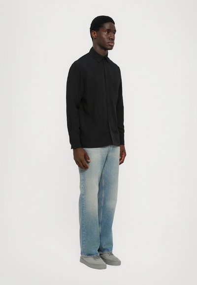 Must have pikendusega must särk, millel on terav krae, kombineeritud helesiniste sirgete pükstega ja hallide kingadega.