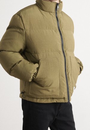 Homme portant une veste matelassée vert olive avec une fermeture éclair frontale et de grandes poches carrées, les mains dans les poches, associée à un pantalon noir.