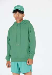 Grüne Kapuzenjacke mit Kordeln und einer Fronttasche, kombiniert mit passenden türkisfarbenen Shorts, die beide das "LES ARCS"-Logo in Weiß tragen.