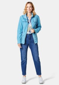 Hellblauem, wasserdichtem Mantel mit verstellbarer Kapuze, Reißverschluss an der Vorderseite und Taschen, über einem gemusterten Hemd getragen, kombiniert mit blauen Jeans und weißen Sneakern.