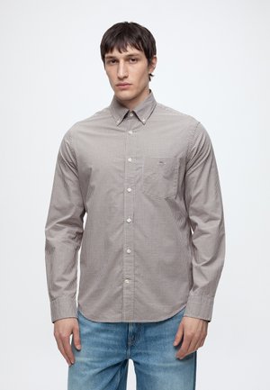 Homme portant une chemise à carreaux marron boutonnée et un jean bleu, debout devant un fond clair uni.