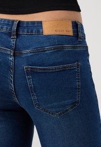 Jeans in denim blu con un'etichetta in pelle che reca la scritta "NOISY MAY" in vita. Presentano cuciture visibili e una tasca posteriore singola.