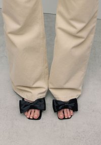 Zwarte satijnen slides met grote strikken en vierkante neuzen, gedragen met lichtbeige wijde broekspijpen die de teennagels onthullen. Luxe texturen en eenvoudig ontwerp.