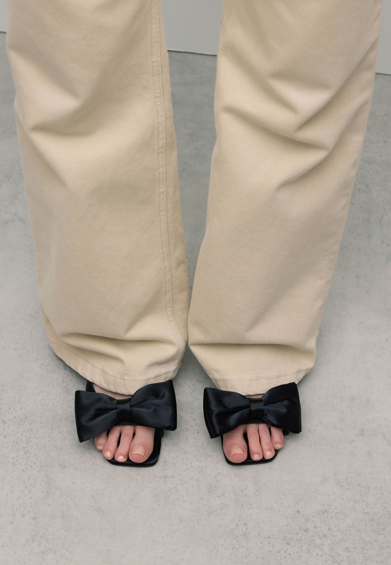 Zwarte satijnen slides met grote strikken en vierkante neuzen, gedragen met lichtbeige wijde broekspijpen die de teennagels onthullen. Luxe texturen en eenvoudig ontwerp.