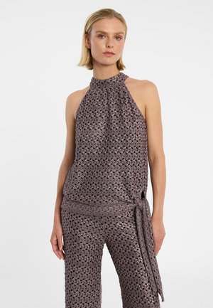 Frau trägt ein ärmelloses, violett gemustertes Neckholder-Top und passende weit geschnittene Hosen mit seitlicher Schleife, stehend vor einem schlichten weißen Hintergrund.