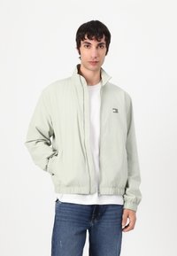 Lys grønn zip-up jakke med elastiske mansjetter og midjebånd, brukt over en hvit T-skjorte. Har et lite logo på brystet.