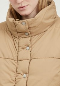 Doudoune beige avec un col haut, ornée de grands boutons pression en argent et de détails cousus sur la texture du tissu.