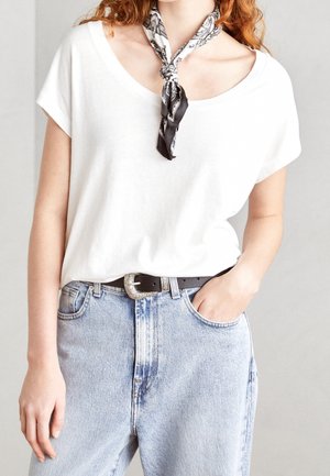 Femme portant un t-shirt blanc à col rond, un jean bleu clair avec une ceinture noire et un foulard noir et blanc à motifs autour du cou, une main dans la poche.